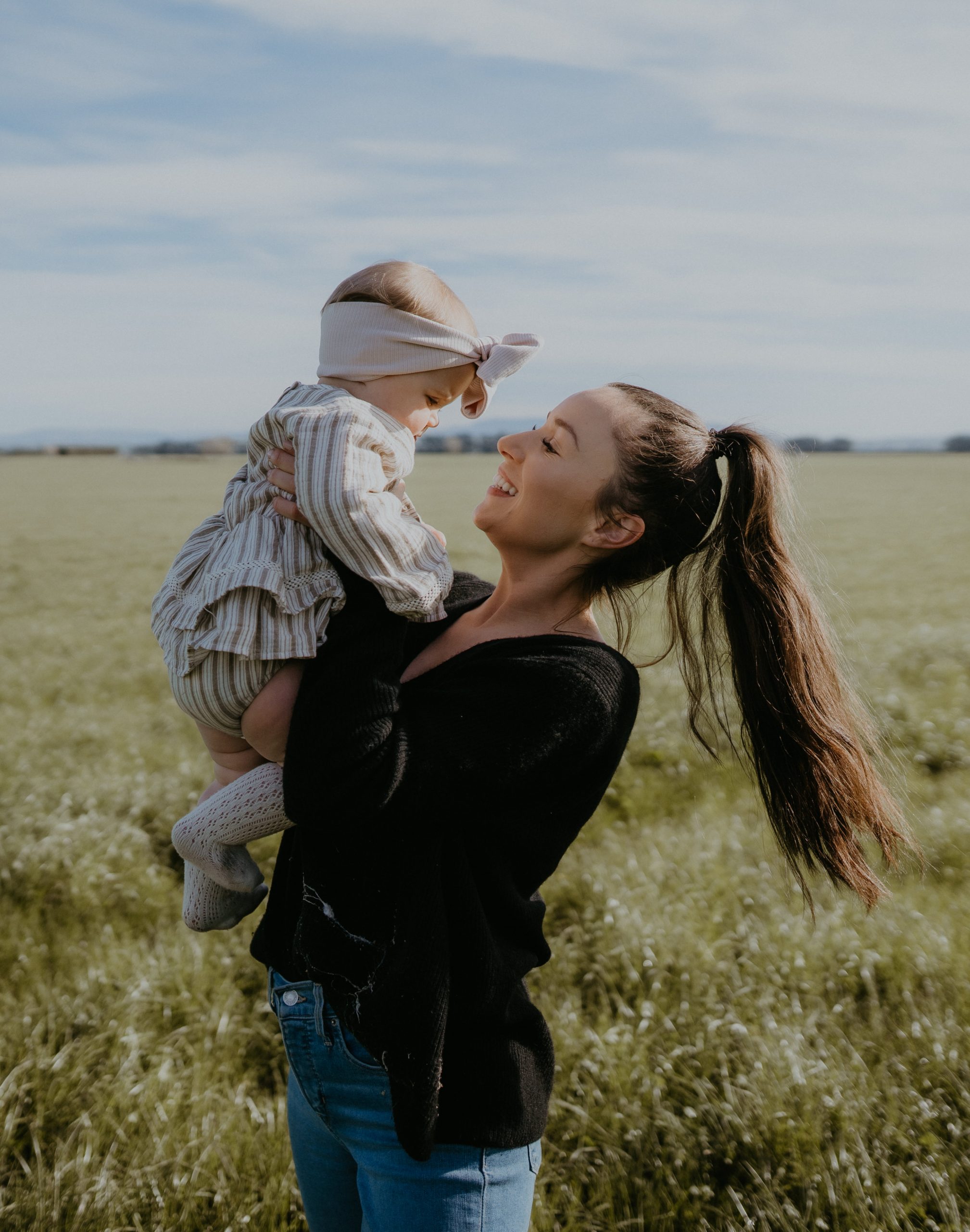 hero-blog-story-tablet Young mother looking into the eyes of her baby whilst holding her up in an open field.